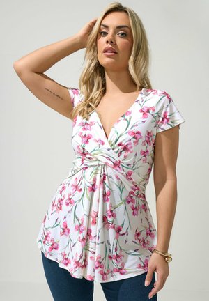 Blonde Frau, die ein weißes Wickeloberteil mit pinken Blumen und blaue Jeans trägt, posiert mit einem Arm hinter dem Kopf erhoben.