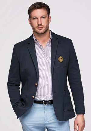 Mand iført navy blazer med guldknapper og emblem, stribet skjorte med knapper, lyseblå bukser, står med den ene hånd i lommen.