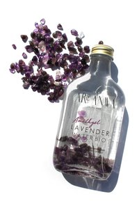 Bouteille en verre clair contenant de l'eau de lavande avec des cristaux d'améthyste, bouchon à vis doré et étiquette blanche avec du texte violet sur un fond lumineux.