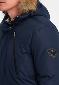 Námořnická modrá zimní bunda s umělou kožešinou na kapuci, kapsou na zip a černou nášivkou na levém rukávu. Hladká textura.