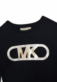 Svart t-shirt med rundad halsringning och en metallisk guldig oval logotyp med "MK" i mitten och ett sammanlänkat kedjemönster runtom.