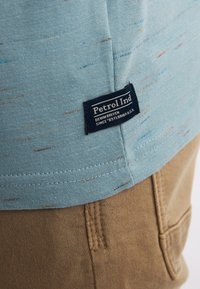 Light blue cotton t-shirt featuring a speckled pattern, with a navy label reading "Petrol Ind" on the hem, paired with brown pants.
