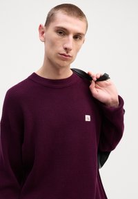 Purple textured sweater with a round neckline and a small logo patch. Model holds a black strap over a shoulder, wearing neutral expression.