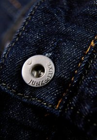 Metalowy guzik z wygrawerowanym napisem "JUNK de LUXE", przymocowany do ciemnoniebieskiego denimu z żółtymi szwami i widocznymi wzorami tekstury.