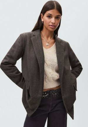 Blazer gris foncé au design ajusté, avec deux poches avant et des revers crantés, porté sur un pull léger en maille et un jean noir avec des accents cloutés.