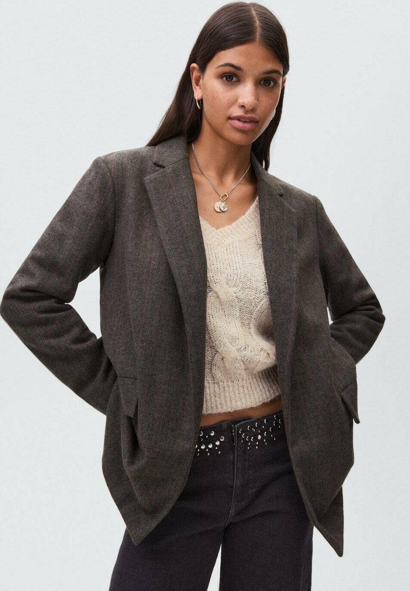 Blazer grigio scuro con design su misura, due tasche frontali e rever a punte, indossato sopra un maglione leggero a maglia e jeans neri con accenti di borchie.