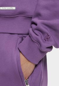 Lila Sweatshirt mit gerippten Bündchen, einer seitlichen Reißverschlusstasche und einem bestickten 'B'-Detail am Ärmel. Weicher Stoff und lockere Passform.