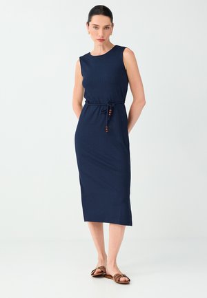Femme aux cheveux foncés en chignon bas, portant une robe midi sans manches bleu marine avec une ceinture nouée à la taille et des sandales plates marron, debout sur un fond blanc.