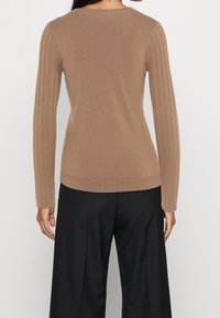 Žena v těsném hnědém žebrovaném svetru s dlouhým rukávem a volných černých kalhotách, zobrazená zezadu na jednobarevném pozadí.