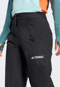 Čierne nepremokavé nohavice so zipsovými vreckami, s logom "TERREX". Vyrobené z odolného materiálu, majú jemnú textúru a mierny lesk.