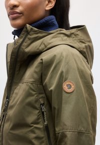 Osoba v olivovo zelenej bunde s kapucňou a okrúhlym logom na hornom rukáve, oblečená cez tmavomodrú fleecovú bundu.