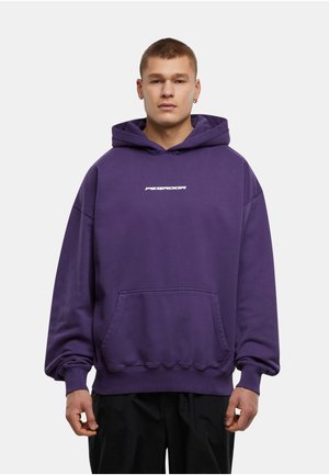 Mladý muž v fialové mikině Pegador s přední kapsou a černými kalhotami stojí před jednoduchým světle šedým pozadím.