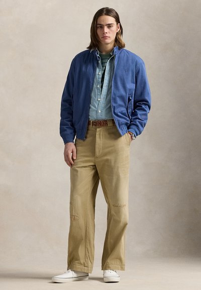Joven de pie con una chaqueta azul sobre una camisa vaquera clara, camiseta verde a rayas, pantalones beige y zapatillas blancas sobre un fondo neutro.