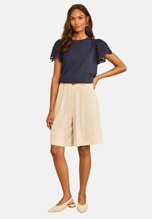 Kvinde iført en navyblå top med blonder korte ærmer, beige knælange shorts, beige lavhælede spidse sko, står med den ene hånd på hoften.