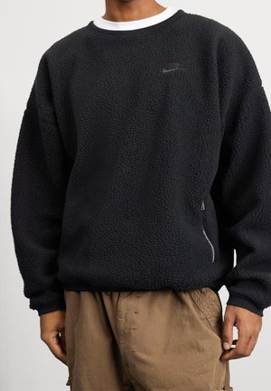Homme portant un sweat Nike noir texturé avec une poche zippée sur le côté droit, un sous-vêtement blanc à col rond, et un pantalon marron.