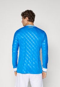 Man facing away wears bright blue long-sleeve sports jersey with subtle diagonal pattern and white shorts against white background.
