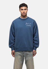 Giovane uomo che indossa una felpa blu con il testo "Good For Nothing" sul lato sinistro del petto e jeans chiari sbiaditi, in piedi contro uno sfondo bianco.