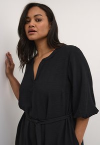 Robe noire ample avec un décolleté en V et des manches courtes, dotée d'une ceinture à nouer à la taille et d'un tissu texturé. Surface lisse avec un léger éclat.