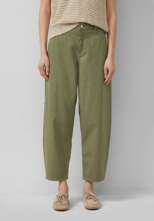 Person trägt olivgrüne, hoch taillierte, weit geschnittene Hosen kombiniert mit beigefarbenem, gestricktem ärmellosem Oberteil und beigefarbenen Schnür-Mokassins.