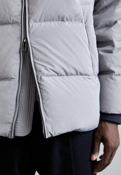 Jachetă puffer gri cu un design matlasat, având un fermoar frontal și o textură netedă. Mânicile și tivul sunt elastice pentru o ajustare confortabilă.