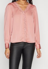 Blouse rose en satin avec un col en V, bordure en dentelle le long de l'encolure et de la patte de boutonnage, et manches longues avec des poignets. Associée à un pantalon noir.