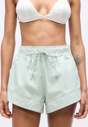Donna che indossa un reggiseno bianco e pantaloncini verde chiaro con coulisse, che mostra un tatuaggio sull'avambraccio sinistro su sfondo neutro.