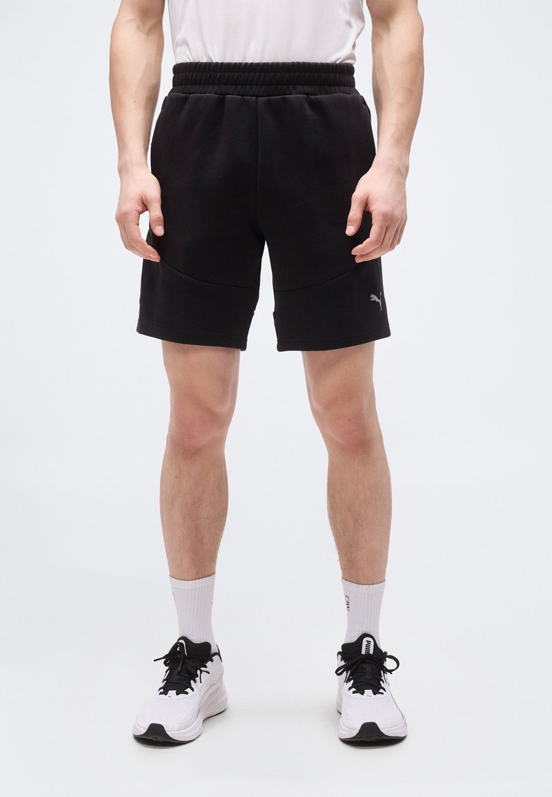 Άνδρας με μαύρο σορτς Puma, λευκές κάλτσες και λευκά αθλητικά παπούτσια Puma, στέκεται μπροστά σε απλό ανοιχτό φόντο.