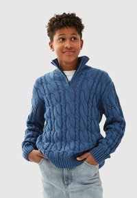 Blå stickad tröja med dragkedja vid halsen, med ett kabelstickat mönster och ribbade ärmslut och fåll, bärs tillsammans med ljusa denimjeans.