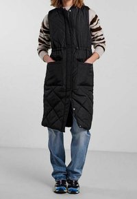 Svart, vadderad lång väst med dragkedja fram, elastisk midja och två framfickor. Bärs över en randig tröja och blå jeans.