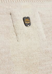 Gebreide trui in lichtbeige met gestructureerde stof, voorzien van een zwart label met gouden tekst. Eenvoudig ontwerp met subtiele stikdetails.