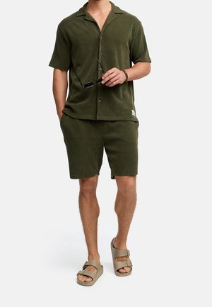 Hombre con camisa de manga corta abotonada y pantalones cortos a juego de color verde oscuro, sosteniendo unas gafas de sol, y con sandalias beige de dos tiras en los pies.