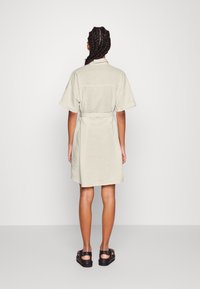 Vestito beige chiaro a maniche corte con cintura, colletto e taglio dritto, rifinito con una sottile texture e lunghezza a metà coscia. Indossato con sandali neri.