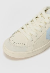 Sneaker with a cream leather upper, light blue accents, perforated toe box, and flat laces. Suede detail at the toe and rubber sole.