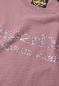 Camiseta de algodón rosa con texto en strass plateados "SuperDry MARAIS. PARIS", con cuello redondo y mangas cortas. Textura suave.