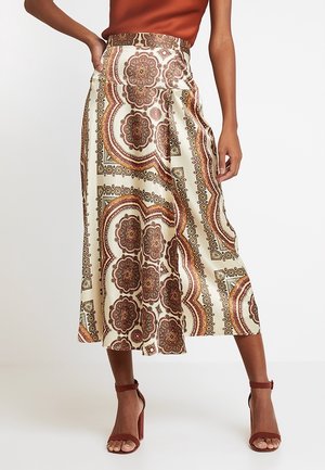 Kobieta w kremowej spódnicy midi z misternymi brązowo-rdzawymi wzorami w kształcie kół, w połączeniu z rdzawymi sandałami na wysokim obcasie z paskiem wokół kostki.