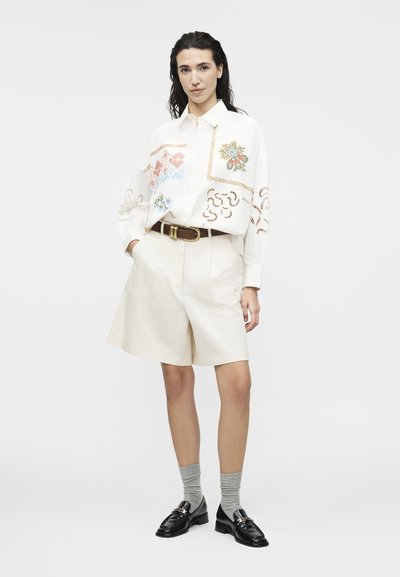 Femme debout portant une chemise blanche à motifs à manches longues, un short beige longueur genou avec une ceinture marron, des chaussettes grises et des mocassins noirs.