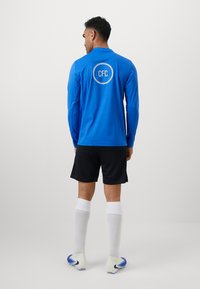 Blå langærmet trøje med "CFC" logo, sorte shorts, hvide sokker og fodboldstøvler med blå accenter. Glat stof og tætsiddende design.