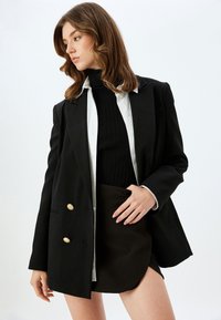 Schwarzes Blazer mit goldenen Knöpfen, über einem schwarzen gerippten Rollkragenpullover getragen, kombiniert mit einem kurzen braunen Rock, vor einem schlichten Hintergrund.