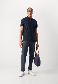 Marinblå polo skjorta med korta ärmar, figurnära design; ihop med mörka byxor och vita sneakers; håller en marinblå tygväska med bruna handtag.