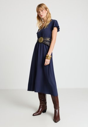 Donna che indossa un vestito midi blu navy, cintura larga intrecciata con grande fibbia rotonda, bracciali d'oro e stivali di pelle marrone fino al ginocchio.