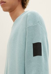 Osoba s světle modrým pleteným svetrem s černým obdélníkovým nášivkem na horní části rukávu, na kterém je napsáno "DENIM TOM TAILOR EST. 2007."