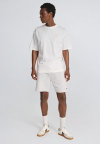 Hvid bomulds-T-shirt med orange logo, parret med lysegrå shorts og cremefarvede sneakers med sorte striber. Glat tekstur, afslappet pasform.