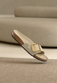 Μπεζ σανδάλι με φαρδιά λουράκι, χρυσή αγκράφα και υφασμένο φελλό στη σόλα. Ο σχεδιασμός έχει ομαλή, γυαλιστερή επιφάνεια.