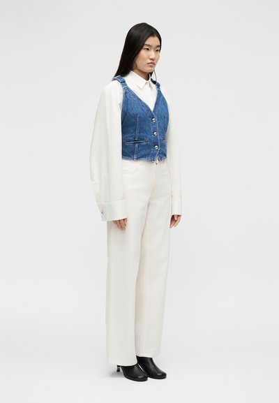 Νέα γυναίκα που φοράει λευκό παντελόνι με ευρεία μπάσκα, λευκό πουκάμισο σε μεγάλο μέγεθος, μπλε τζιν γιλέκο και μαύρα αστραγάλια με τακούνι, στέκεται σε απλό φόντο.