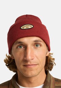 Berretto a costine rosso con una toppa ovale color crema che presenta testi e grafiche neri. Accentuato da una finitura liscia e testurizzata.