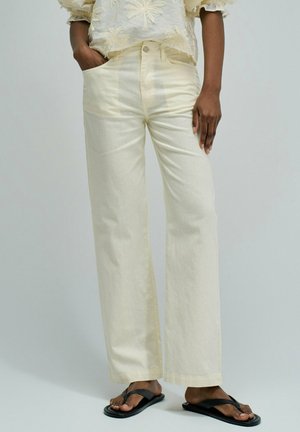 Personne portant un pantalon droit couleur crème, un haut texturé crème et des tongs noires, debout avec une main dans la poche.