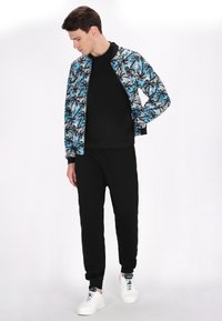 Bomberjack in blauw en zwart met een tropisch bladerenpatroon, gedragen over een zwart shirt en gecombineerd met zwarte joggers en witte sneakers.