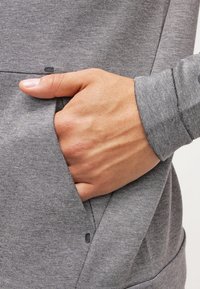Šedý svetr se boční kapsou, se záběrem na ruku odpočívající uvnitř kapsy. Materiál je hladký s jemnou strukturou.