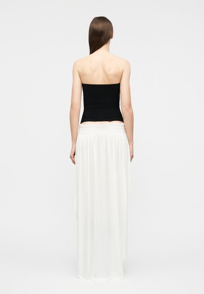 Γυναίκα με μακριά καστανά μαλλιά στέκεται με την πλάτη προς την κάμερα, φοράει μαύρο τοπ χωρίς τιράντες και μακριά λευκή φούστα που κυλά, μπροστά σε απλό φόντο.