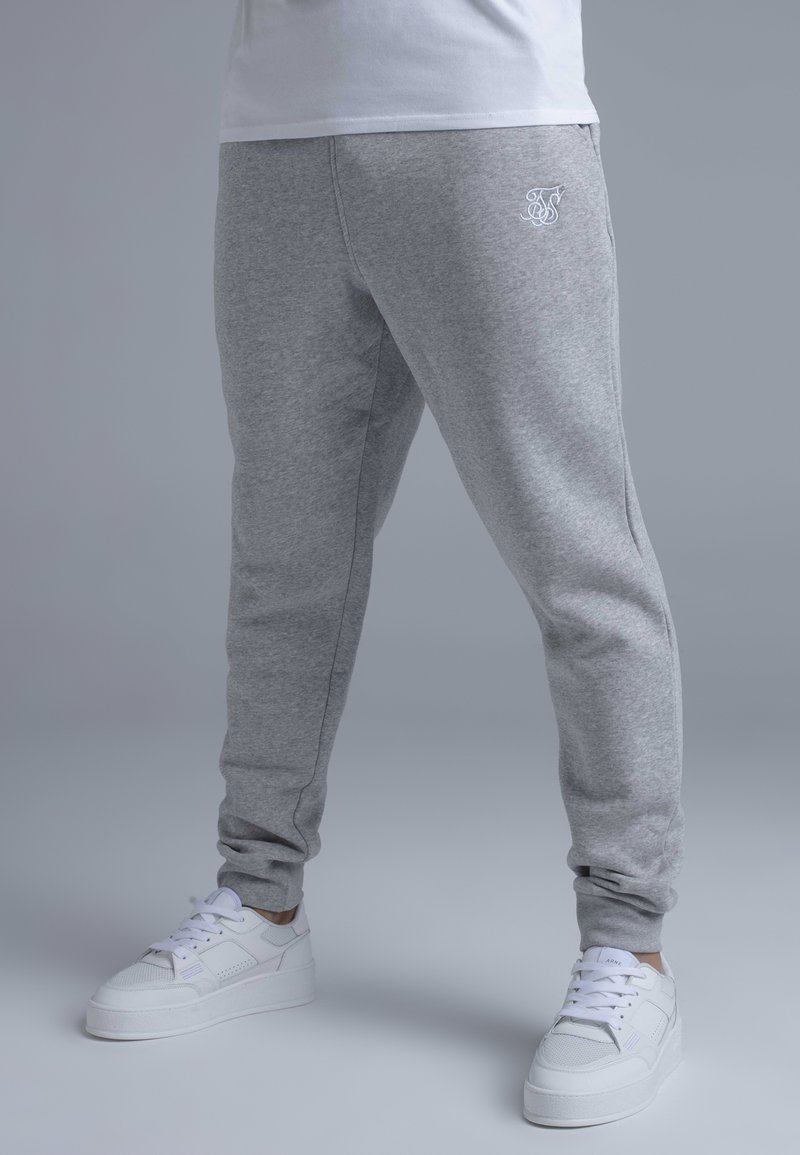 Grey sweatpants made of soft fabric with a tapered fit, featuring embroidered logo detail and elastic cuffs. Worn with white sneakers.